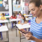 schoolteacher-using-digital-tablet-at-school-in-classroom-at-school-with-school-kids-in-background.jpg